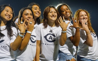 A group of UC Davis Women's Gymnastics team members pose with plain white T-shirts with the UC Davis Gymnastics logo while flashing football rings on their hands at night at a football game.