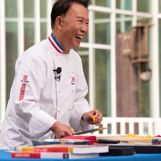 Chef Martin Yan wearing a white chef smock and smiling while presenting at a cooking demonstration in the courtyard of the UC Davis Library.